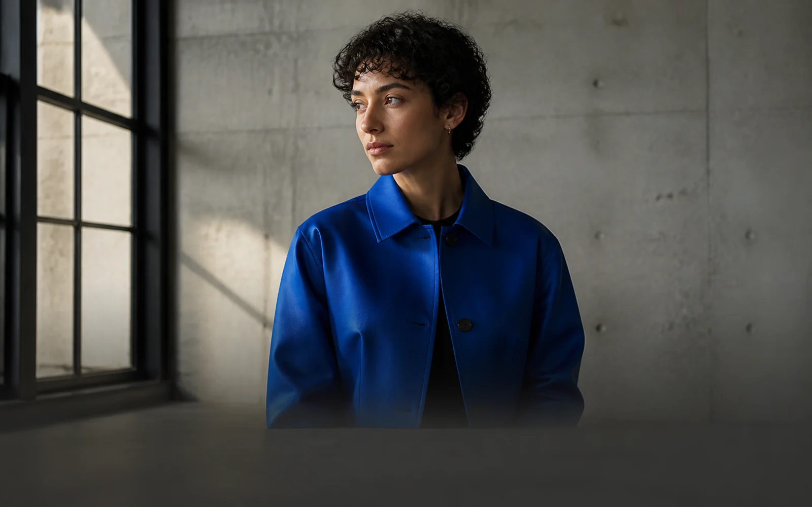 Short-haired fashion model wearing a structured cobalt-blue jacket in a minimal concrete studio.