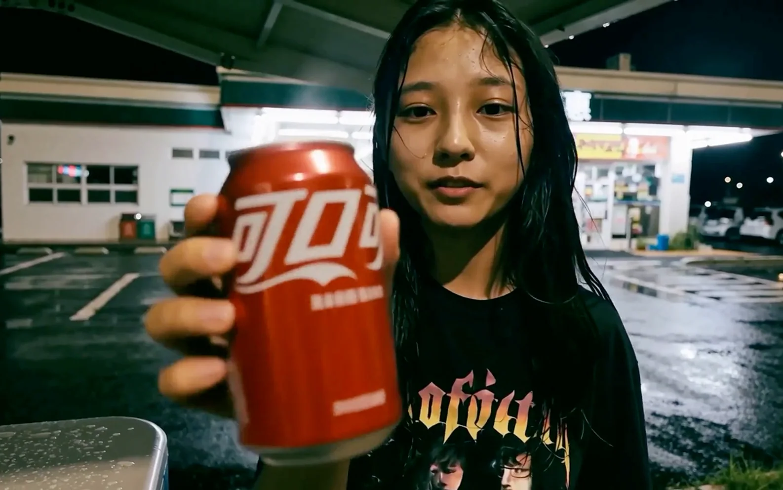 Young woman holding a red soda can toward a handheld camera outside a brightly lit convenience store after rain.