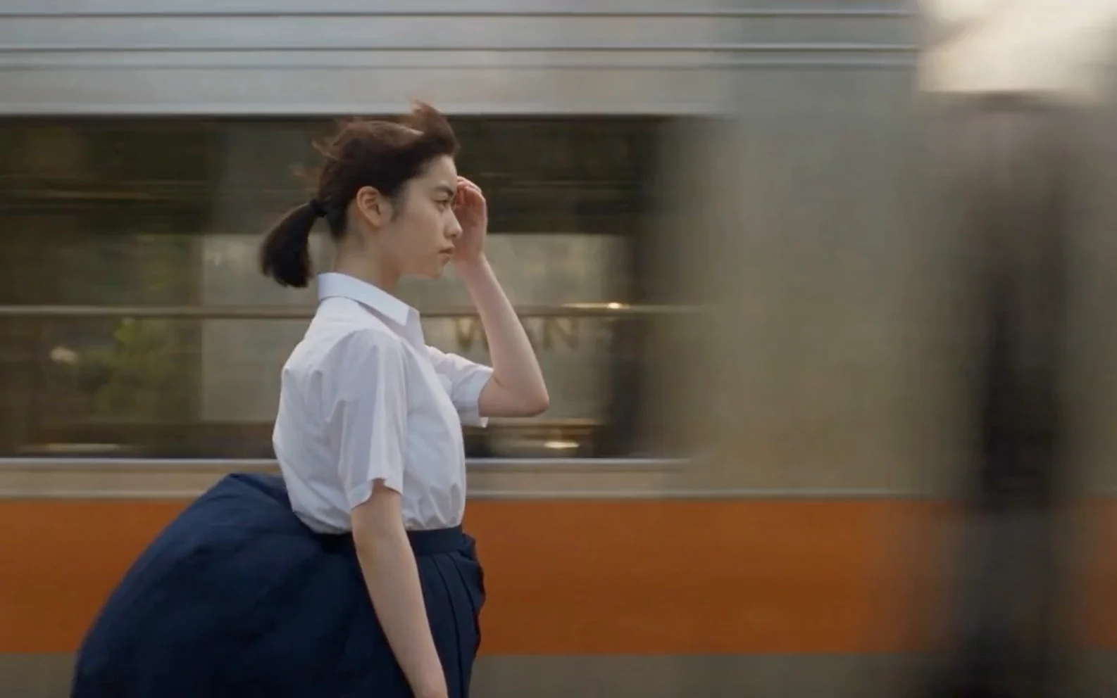 Young East Asian woman in a white blouse walking beside a moving orange train while holding a small red fruit near her face.